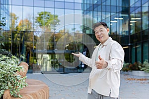 A young Asian man, a businessman, a freelancer, is standing outside an office, a skyscraper, and points to the entrance