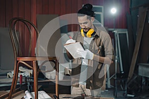 Young Asian man Carpenter uses a tape measure to measure chair on the workbench in woodcraft carpentry workshop.