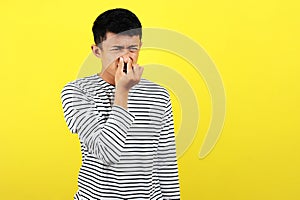 Young Asian man close his nose for bad smell, isolated on yellow