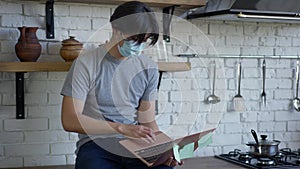 Young Asian man in Covid face mask texting typing on laptop keyboard sitting on kitchen countertop. Portrait of