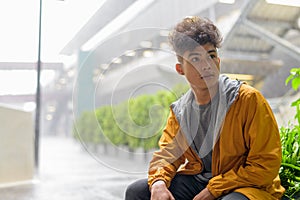 Young Asian man with curly hair thinking and sitting in the city outdoors with rain