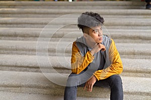 Young Asian man with curly hair thinking and sitting on staircase in the city