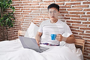 Young asian man drinking coffee sitting on the bed scared and amazed with open mouth for surprise, disbelief face