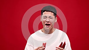 Young asian man eating red hot chilli peppers against red studio background. Crying, spicy hot taste. Grimace face