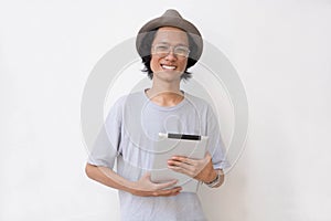 Young asian man with fedora hat and glasses using tablet and smiling at camera holding a tablet pc