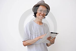 A young asian man with fedora hat and glasses using tablet and smiling at camera while holding tablet pc and using it