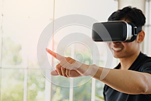 Young Asian man getting experience with VR virtual reality headset or 3D glasses pointing with his finger in living room at home.