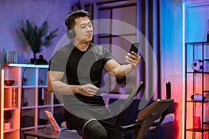 Young asian man with headphones doing exercises on stationary bicycle at home or fitness center.