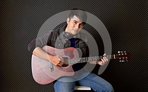 Young asian man with headphones playing an acoustic guitar in front of black soundproofing wall. Musician producing music in