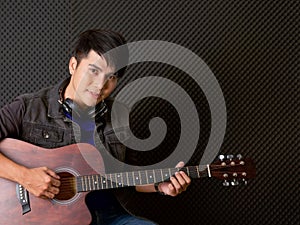 Young asian man with headphones playing an acoustic guitar in front of black soundproofing walls. Musicians producing music in