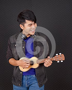 Young asian man with headphones playing an Ukulele guitar in front of black soundproofing walls. Musicians producing music in