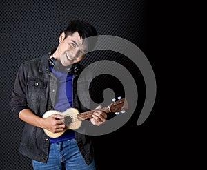 Young asian man with headphones playing an Ukulele guitar in front of black soundproofing walls. Musicians producing music in