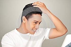 Young Asian man is lokking in mirror and touching his hair on white background