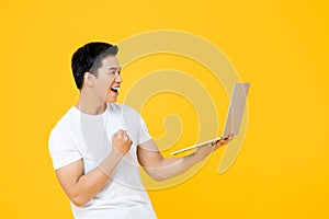 Young Asian man looking at laptop computer and raising his fist doing yes gesture isolated on yellow background