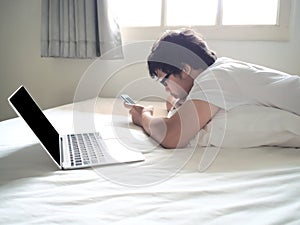 Young Asian man with mobile smart phone lying down on the bed in morning. Internet of things concept