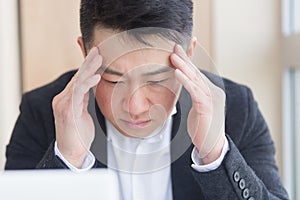 Young asian man, office worker sitting at workplace holding hands massages forehead and head with severe pain. Male at work
