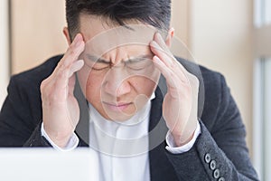 Young asian man, office worker sitting at workplace holding hands massages forehead and head with severe pain. Male at work