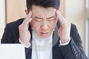 Young asian man, office worker sitting at workplace holding hands massages forehead and head with severe pain. Male at work