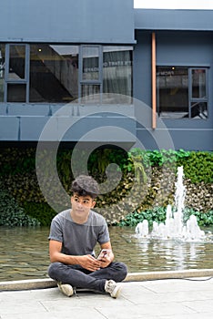 Young Asian man with phone thinking and sitting outside modern building