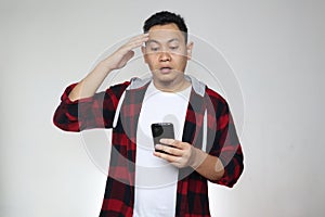 Young Asian man reading texting chatting  on his phone, shocked worried surprised expression, over grey