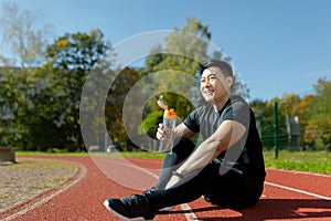 Young Asian man resting after jogging, competition, sitting on treadmill in stadium and drinking water from bottle
