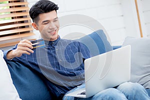 Young asian man sit on sofa using laptop computer shopping online with credit card buying to internet.