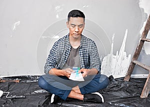 A young Asian man sits on the floor choosing paint colour from swatches home DIY