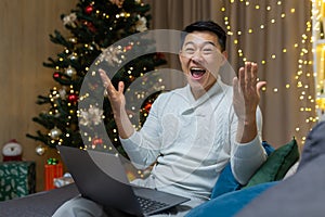 A young Asian man sits at home, holds a laptop, rejoices at the Christmas win in the raffle