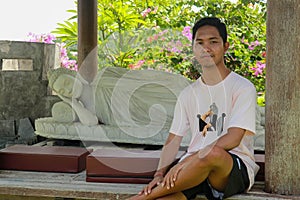 A young Asian man sits by a white Buddha statue lying in the Brahma Vihara Arama Temple. Statue of Sleeping Buddha at