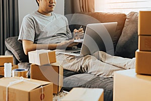 Young asian man sitting on sofa looking on computer laptop and writing order list on paper, SME concept