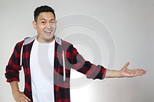 Young Asian man smiling, showing and pointing something on his side, against white wall with copy space