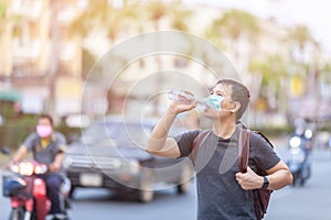 A young asian man standing drinks water with bottle of water in his hands, waiting for the car at road, travel.