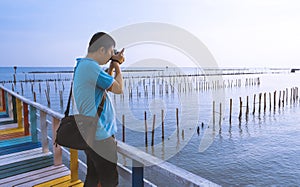 Young Asian man taking picture with digital camera on the old multicolored wooden bridge at sea viewpoint