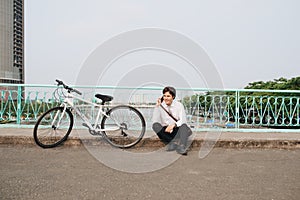 Young asian man talking by mobile phone while sitting on the ground near the bike, outdoors. Wearing stylish clothes.