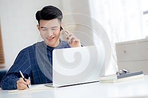 Young asian man talking phone and work from home with laptop computer, freelance male writing on notebook.