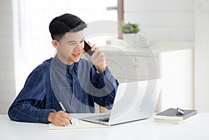 Young asian man talking phone and work from home with laptop computer, freelance male writing on notebook.