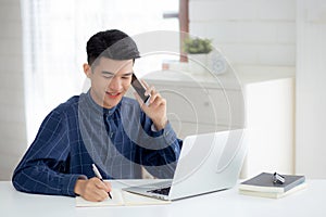 Young asian man talking phone and work from home with laptop computer, freelance male writing on notebook and speak.