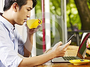 Young asian man using cellphone and laptop while drinking coffee