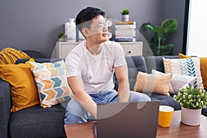 Young asian man using laptop at home sitting on the sofa looking away to side with smile on face, natural expression