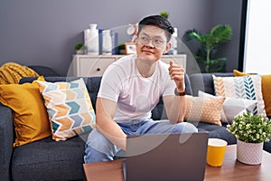 Young asian man using laptop at home sitting on the sofa smiling looking to the side and staring away thinking