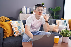 Young asian man using laptop at home sitting on the sofa surprised pointing with hand finger to the side, open mouth amazed