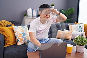 Young asian man using laptop at home sitting on the sofa very happy and smiling looking far away with hand over head