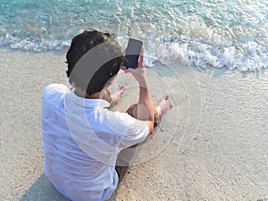 Young Asian man using mobile smart phone on the sandy beach. Summer vacations concept.