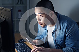 Young Asian man using smart phone and computer until late night, he is working or studying hard to reach his ambition. Student or