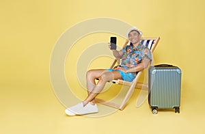 Young asian man wear summer clothes sit in deckchair showing mockup mobile cell phone app booking hotel isolated on yellow