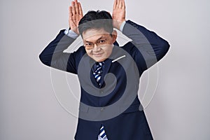 Young asian man wearing business suit and tie doing bunny ears gesture with hands palms looking cynical and skeptical