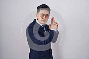 Young asian man wearing business suit and tie holding symbolic gun with hand gesture, playing killing shooting weapons, angry face