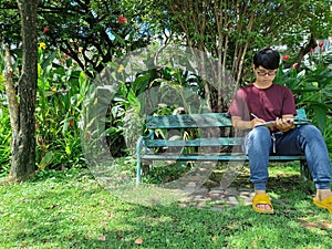 Young Asian man wearing glasses with black hair holding a book on a bench. happy and relax. What are thinking in the summer park