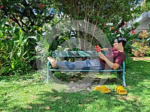 Young Asian man wearing glasses with black hair holding a book sitting with legs stretched on a bench. happy and relax. What are