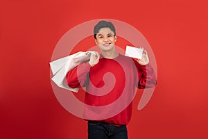 Young Asian man wearing a red sweater is holding a credit card and shopping bags on a red background
